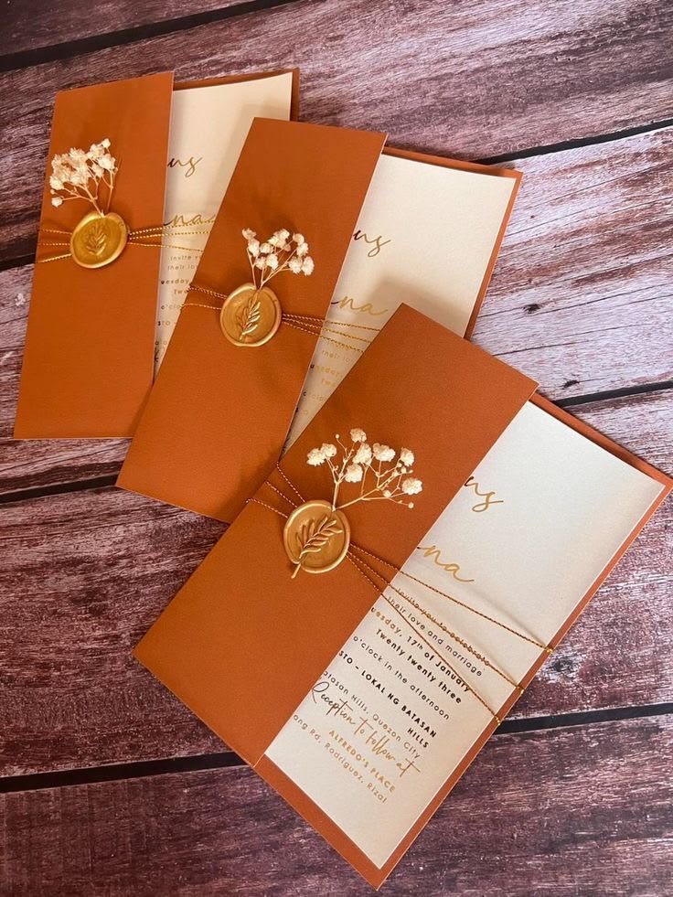 Terracotta wedding invitations with cream inserts, gold cord, dried flowers, and botanical wax seals on a wooden surface.
