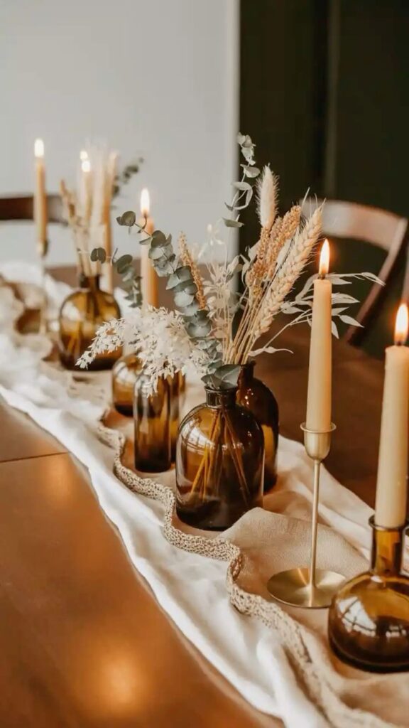 Amber glass bottles filled with dried grasses and eucalyptus beside tall taper candles on a neutral table runner.