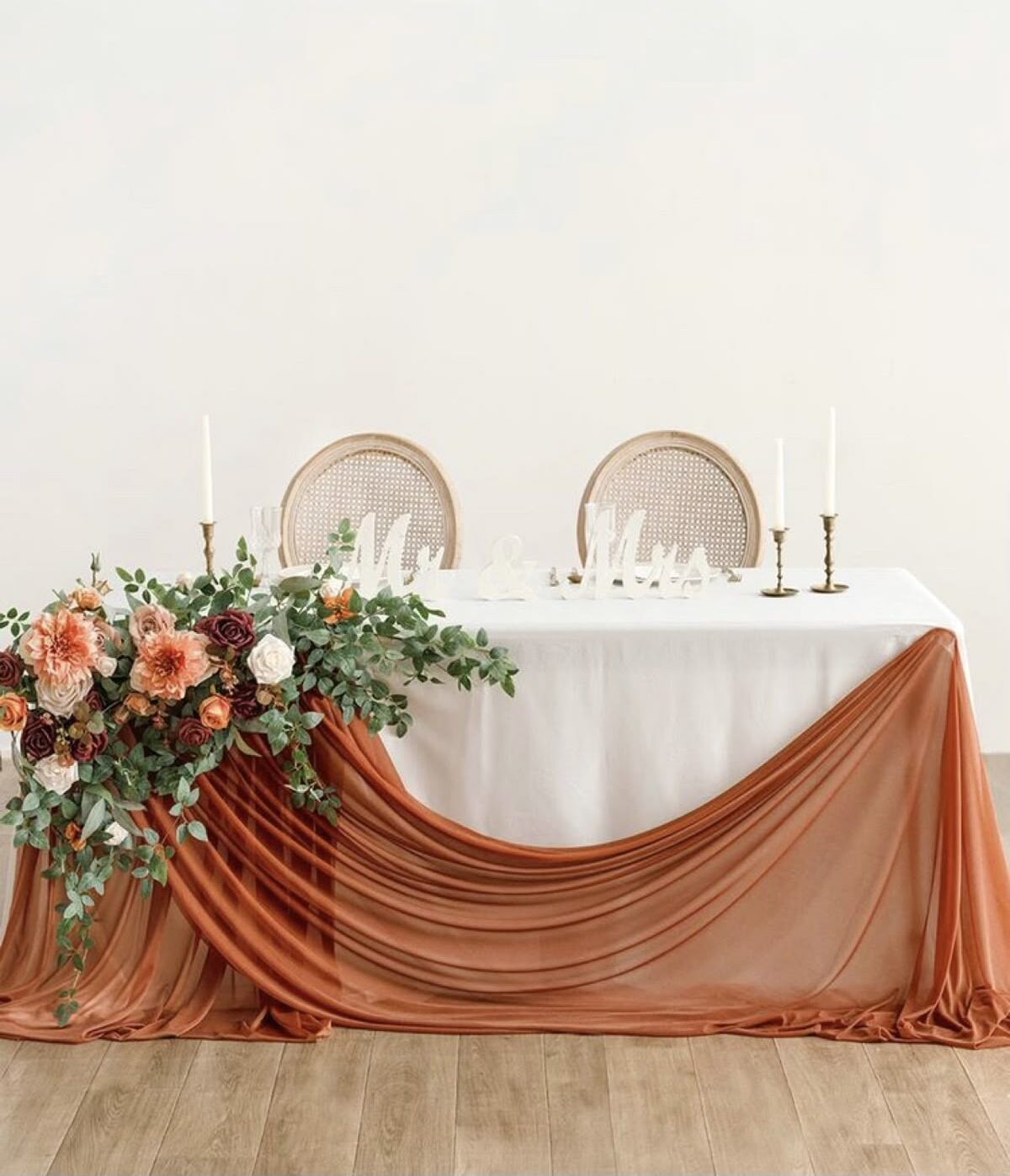 Wedding table with white linen, terracotta draping, cream and rust floral arrangement, and brass candlesticks.