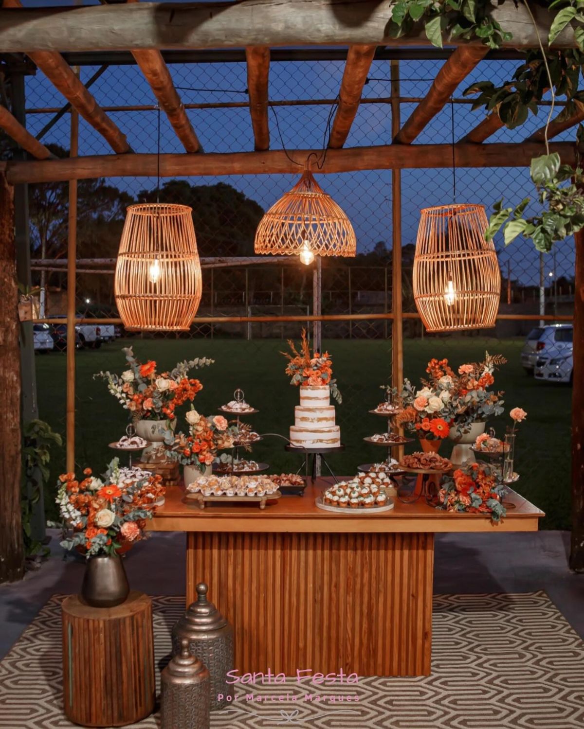Rustic wedding dessert table with tiered cake, cupcakes, orange floral arrangements, and woven pendant lights.
