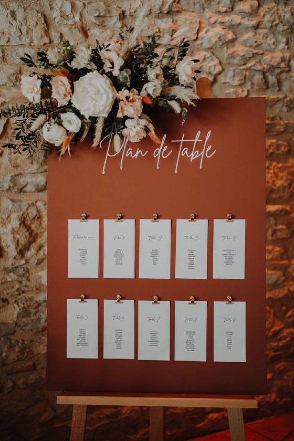 Terracotta wedding seating chart with cream place cards clipped to a board beneath an autumn floral arrangement.