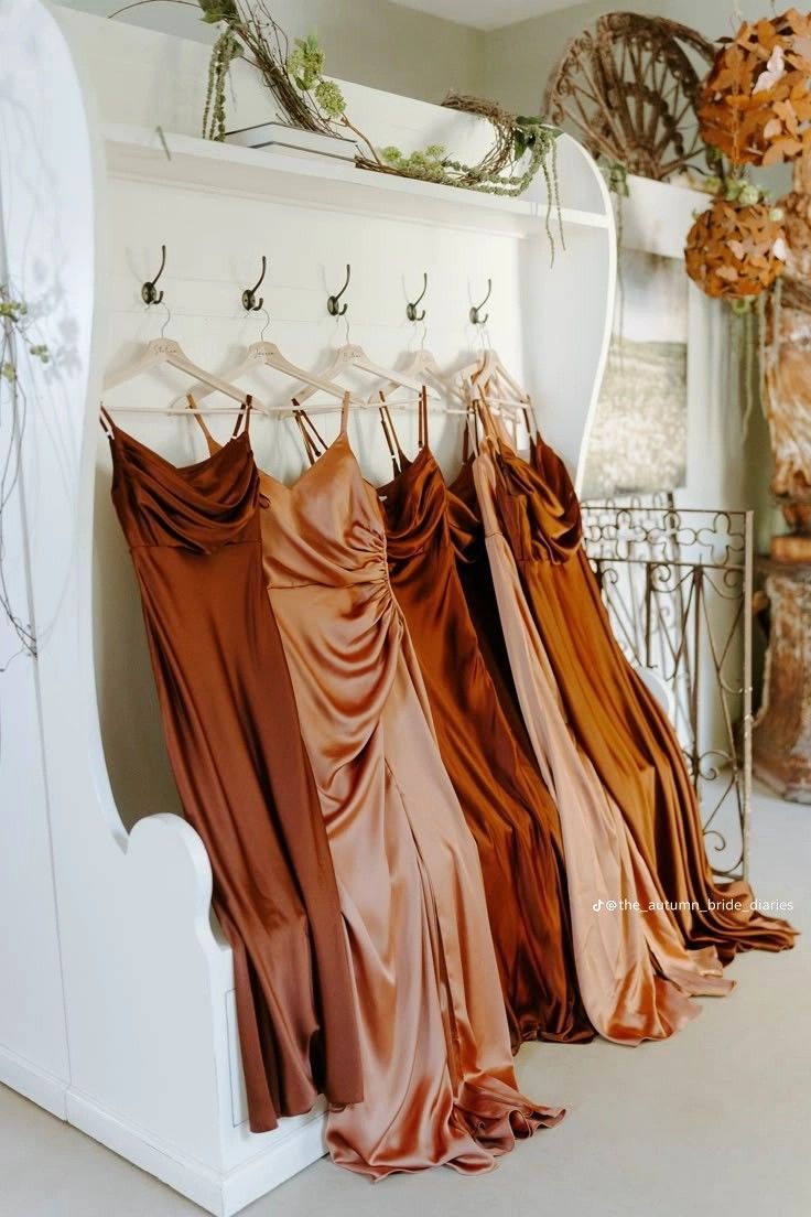 Bridesmaid dresses in copper, caramel, burnt orange, and rust satin hanging together on wooden hangers.
