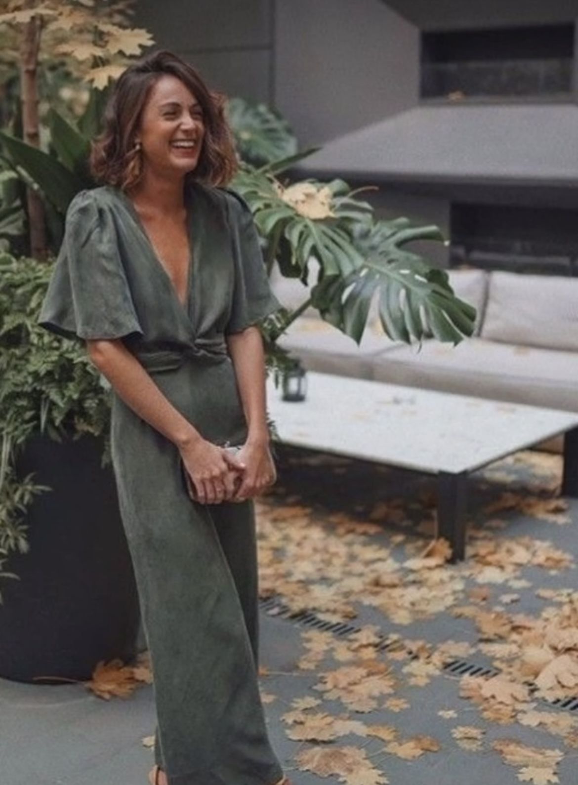 Woman wearing a sage green wrap midi dress at an outdoor setting with fall leaves
